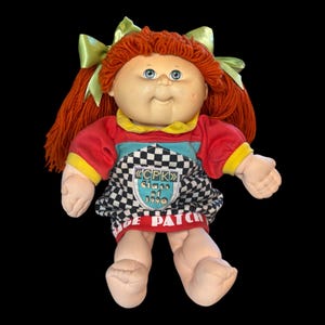May include: A vintage Cabbage Patch Kids doll with red yarn hair and blue eyes. The doll wears a red, blue, and black checkered dress with the text "CPK Class of 1990" and green bows in its hair.