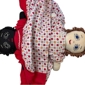 May include: Two handmade cloth dolls. One doll has black skin, black hair, and is wearing a red dress. The other doll has light skin, brown hair, and is wearing a patterned dress with red, pink, and orange circles. The dolls are partially covered by a patterned blanket.