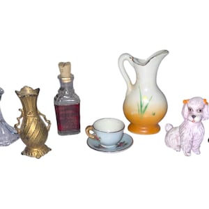 May include: A collection of miniature decorative items including a glass decanter, a gold-colored vase, a porcelain pitcher, a teacup and saucer, a porcelain dog figurine, and a glass oil lamp.