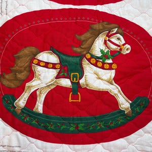 May include: A Christmas-themed fabric panel featuring a white rocking horse with a brown mane and tail, adorned with a green saddle and gold accents. The horse is set against a red oval background with a quilted pattern.