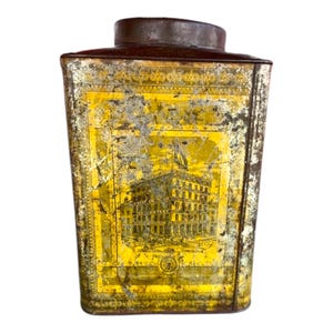 May include: Vintage metal tea tin with a yellow and gold design. The tin features a building with a large window and a sign that reads "The Great Atlantic & Pacific Tea Co."