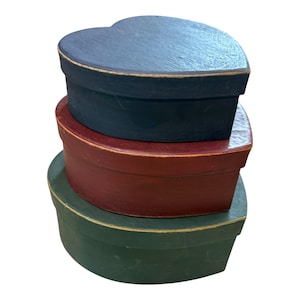 May include: Three heart-shaped decorative boxes stacked on top of each other. The top box is navy blue, the middle box is burgundy, and the bottom box is green. The boxes have a rustic, painted finish.