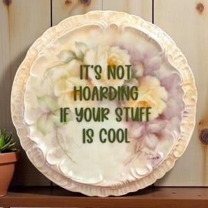 May include: A decorative, round plate with a floral design in yellow, pink, and green. The plate has a scalloped edge and the text "IT'S NOT HOARDING IF YOUR STUFF IS COOL" in green lettering.