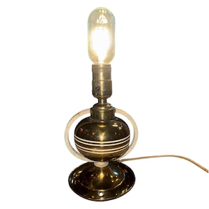 May include: A vintage brass table lamp with a bulb. The lamp has a round base and a spherical body with white stripes. A light-colored, curved accent surrounds the body. The bulb is elongated and lit, with a visible filament.