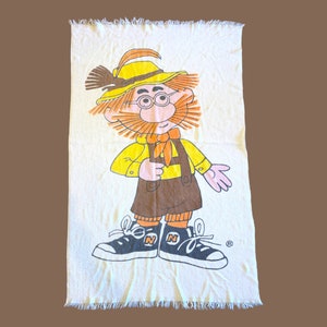 May include: A white towel with a cartoon illustration of a man wearing a yellow shirt, brown pants, a yellow hat, and brown shoes. The man has a red beard and mustache and is holding a broom. The towel has a fringed edge.