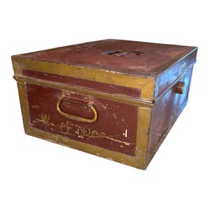 May include: A rectangular, vintage metal box with a hinged lid. The box is painted burgundy with gold trim and a gold handle. The box has a slot on the top and text on the front. The box is likely used for storage.