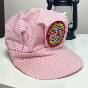 May include: Pink and white striped cap with a rounded crown and a short brim. The cap features a circular patch with the words "Manitou and Cog Wheel Route Pike's Peak Railway" in a gold and red design. The cap is made of a striped fabric.