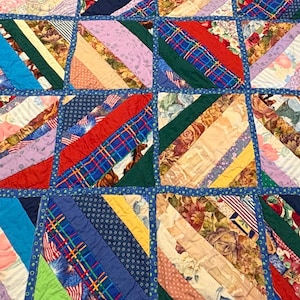 May include: A patchwork quilt with a variety of fabrics in a geometric pattern. The quilt features diagonal stripes in shades of blue, red, green, purple, and beige. The quilt is bordered with a blue fabric.