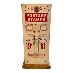 May include: Vintage white metal postage stamp dispenser with red lettering. The dispenser has a slot for inserting coins and a compartment for dispensing stamps. The text on the dispenser reads "U.S. Postage Stamps in Sanitary Folders" and "The Stampmaster 10¢".