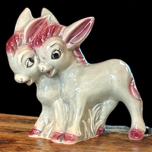 May include: A ceramic figurine of a two-headed donkey with pink ears and a gray body. The donkey is standing on a wooden surface.