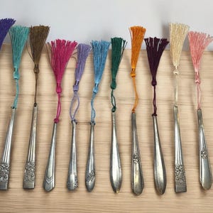 May include: A set of 14 silver bookmark spoons with colorful tassel charms. The tassels are in various colors, including purple, teal, brown, pink, lavender, blue, green, orange, burgundy, cream, peach, and light green.