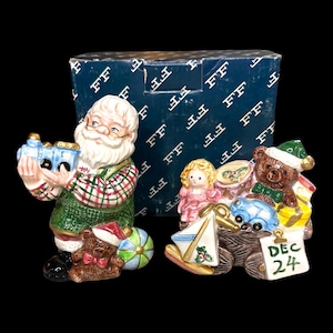 May include: Christmas figurines featuring Santa Claus holding a toy train and a second figurine with a teddy bear, doll, and gifts. The figurines are colorful with intricate details, and the box behind them has a blue background with the letter "F" repeated.