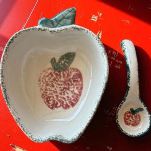 May include: Apple-shaped ceramic serving bowl and spoon set. The bowl and spoon are white with a red and green apple design. The rim of the bowl and spoon are trimmed with green. The set is on a red surface.