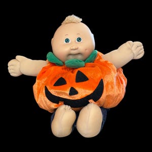 May include: A vintage Cabbage Patch Kid doll dressed in an orange pumpkin costume with a black jack-o'-lantern face. The doll has a fair complexion, blue eyes, and a tuft of blonde hair. It is wearing blue denim pants.