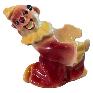 May include: A ceramic clown planter with a red and yellow costume. The clown is kneeling and has a wide smile with a red nose. The planter has a small opening at the top for planting.