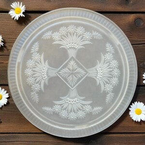 May include: A clear, round decorative plate with an embossed floral design. The plate features a central diamond shape and surrounding floral patterns. White daisies and a pink flower are scattered on a dark brown wooden surface.