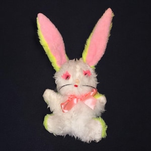 May include: A vintage plush bunny toy with long ears. The bunny is white with pink and yellow accents on the ears and feet. It has pink eye details, a red nose, black whiskers, and a pink ribbon bow. The toy is on a dark background.