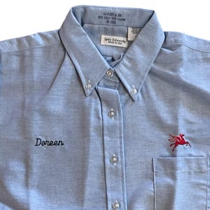 May include: A light blue long-sleeved button-down shirt with a black embroidered name "Doreen" on the left chest and a red embroidered winged horse logo on the right chest. The shirt has a pocket on the right side.