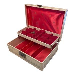 May include: A two-tiered jewelry box with a beige exterior and red velvet lining. The top tier has multiple compartments and the bottom tier has a single long compartment. The box has a keyhole and a small key is visible in the top tier.