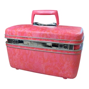May include: A rectangular pink vanity case with a textured surface and a solid pink top handle. The case features a horizontal silver-toned metal band across the center, which includes two circular key locks and a central rectangular latch mechanism.