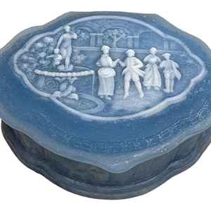 May include: A blue trinket box with a detailed relief scene on the lid. The relief depicts figures in a garden setting, including a fountain and several people. The box has a scalloped edge and a dark base.