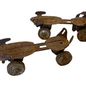 May include: A pair of vintage rusty roller skates with black straps. The skates have four wheels each and are made of metal.