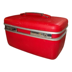 May include: A vintage red travel suitcase with a textured surface. The rectangular case has a silver-colored metal trim and handle. A keyhole is visible on the front. The suitcase is likely from the mid-20th century.