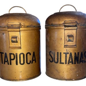 May include: Two antique, cylindrical metal canisters with lids and handles. One reads "TAPIOCA" in black letters, the other "SULTANAS". The canisters have a gold finish with visible wear and tear.