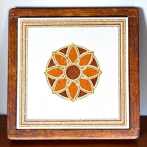 May include: Square wooden trivet with a white tile insert. The tile features a central floral design in shades of brown and orange, surrounded by a gold border. The wooden frame is a warm brown color.