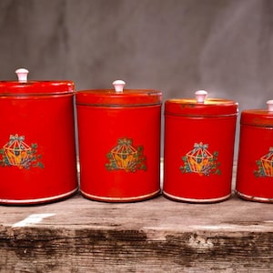 May include: Set of four vintage red metal kitchen canisters with white lids. Each canister features a decorative illustration of a bird cage with fruit and foliage. The canisters are arranged on a weathered wooden surface.