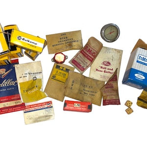 May include: A collection of vintage automotive parts and packaging, including boxes for Cadillac, Delco, Balkamp, and FoMoCo, as well as a small gauge, a red plastic part, and a pair of dice.