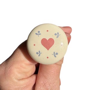 May include: A round, cream-colored knob with a pink heart in the center. Small blue floral designs and red dots surround the heart. The knob is held in a hand.
