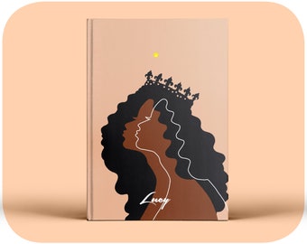 Könnte beinhalten: Ein Hardcover-Buch mit hellpfirsichfarbenem Einband, das eine schwarze Silhouette einer Person mit langem lockigem Haar und einer Krone zeigt. Eine weiße Linienzeichnung umreißt das Profil und der Name Lucy steht in weißer Schrift.