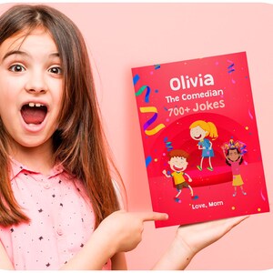 Op de afbeelding: Een jong meisje houdt een rood boek vast met de titel "Olivia The Comedian 700+ Jokes" en de woorden "Love, Mom" onderaan. De kaft van het boek bevat cartoonillustraties van kinderen op een podium met confetti.