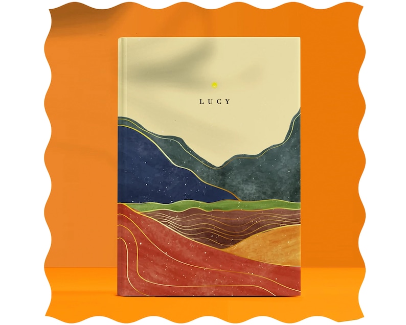 May include: A cream-colored journal with the word "LUCY" printed in gold lettering. The cover features a watercolor-style landscape with blue, green, and brown hills and gold accents.