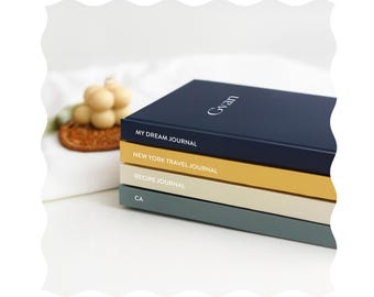 Op de afbeelding: Een stapel van vier dagboeken met verschillende gekleurde omslagen. Het bovenste dagboek is donkerblauw met de tekst "MY DREAM JOURNAL" en "Gvan" in wit. Het tweede dagboek is geel met de tekst "NEW YORK TRAVEL JOURNAL" in wit. Het derde dagboek is crèmekleurig met de tekst "RECIPE JOURNAL" in wit. Het onderste dagboek is grijs met de tekst "CA" in wit.