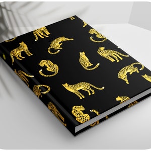 Könnte beinhalten: Ein schwarzes Notizbuch mit einem goldenen Leopardenmuster. Das Notizbuch hat einen weißen Rücken und eine weiße Kante.