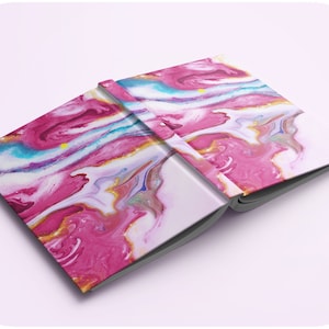 May include: A pink and white notebook with a marbled cover. The cover has swirls of pink, white, blue, and gold paint.