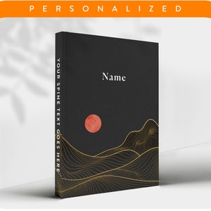 Op de afbeelding: Een zwart notitieboek met een gouden en wit ontwerp van bergen en een rode zon. De rugtekst luidt "YOUR SPINE TEXT GOES HERE". De voorkant heeft het woord "Name" in wit gedrukt.