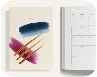 Puede incluir: Un diario de color crema con un diseño de acuarela con detalles azules, rosas y dorados. La portada tiene la palabra "GVAN" impresa en naranja. El diario está abierto en una página de calendario de febrero.