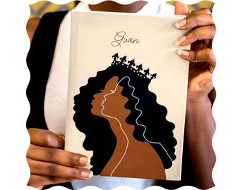 May include: A beige journal with a black and brown illustration of a woman wearing a crown. The journal has the name "Guan" written on the front.