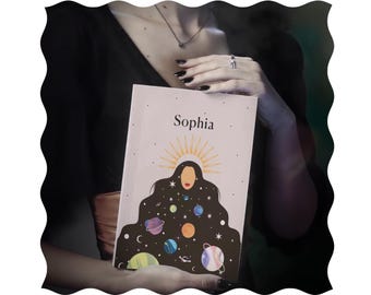 Op de afbeelding: Een roze notitieboek met de naam "Sophia" gedrukt op de voorkant. De omslag heeft een zwart silhouet van een vrouw met een halo van sterren en planeten rond haar hoofd.