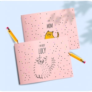 Puede incluir: Dos libros de actividades de puntos a conectar de color rosa con temática de gatos. El libro de arriba dice "Love Mom" y el libro de abajo dice "I'm Busy Lucy".