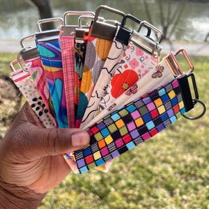 May include: A collection of colorful fabric keychains with different patterns and designs. The keychains are made of fabric and have metal rings. The keychains are held in a hand.