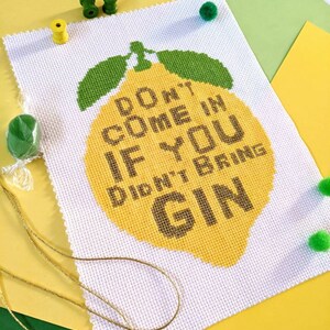 Könnte beinhalten: Ein Kreuzstich-Kunstwerk auf weißem Stoff zeigt eine große gelbe Zitrone mit grünen Blättern. Der Text "DON'T COME IN IF YOU DIDN'T BRING GIN" ist in Braun gestickt. Das Kunstwerk ist von gelbem, grünem und weißem Papier umgeben.