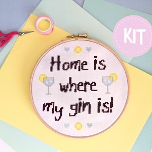 以下が含まれることがあります： 白い背景に黒い文字で「Home is where my gin is!」と書かれたクロスステッチ刺繍キット。ジンとトニックのイラストが2つあります。キットは木製の刺繍枠に入っています。