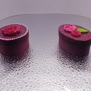 May include: Two small round boxes with floral designs. One box is round and the other is oval. Both boxes are a deep pink color with a textured surface. The round box has a single pink rose with a pearl border. The oval box has three pink roses and a green leaf.