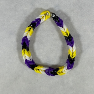 以下が含まれることがあります： 紫、黒、白のゴムバンドで作られたブレスレット。ブレスレットは、複数のゴムバンドの糸が織り込まれて作られています。