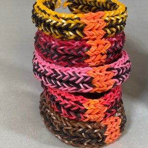 May include: A stack of five colourful rubber band bracelets. The bracelets are made with black, yellow, pink, red, and orange rubber bands. The bracelets are woven together in a herringbone pattern.