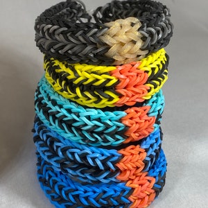 May include: A stack of six colorful rubber band bracelets. The bracelets are made with black and a variety of bright colors, including yellow, blue, and orange. The bracelets are stacked on top of each other, with the top bracelet being a sparkly gold color.